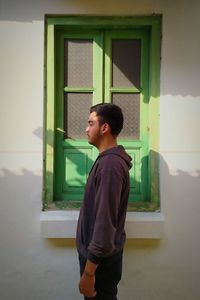 Side view of young man looking away while standing against window