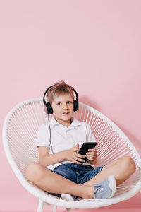 Portrait of boy listening music while using phone on chair against colored background
