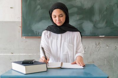 Teacher wearing traditional clothing writing on book while sitting against blackboard in classroom