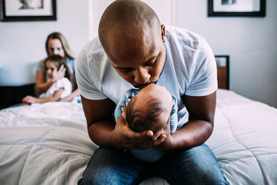 Dad kissing newborn on bed with mom and daughter in back ground