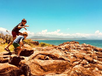 Boys playing on rock by sea against sky