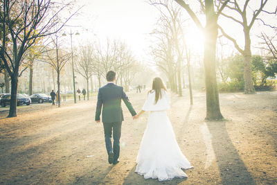 Rear view of bride and bridegroom holding hands while walking amidst bare trees during sunset
