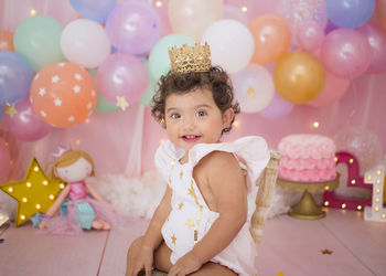 Cute baby girl with balloons and birthday cake sitting at home