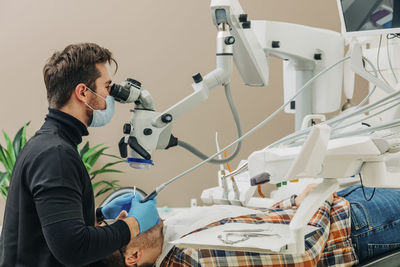 Young dentist examining patient's teeth through microscope in clinic