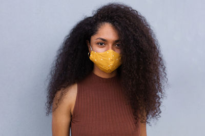 Positive african american female with curly hair wearing yellow protective mask standing near urban building in city during coronavirus pandemic and looking at camera