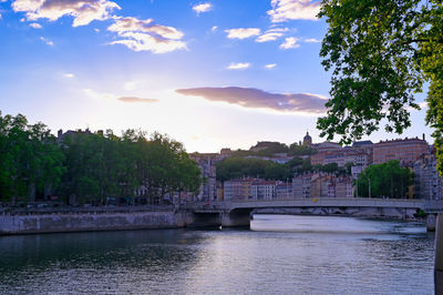 Bridge over river by buildings against sky during sunset