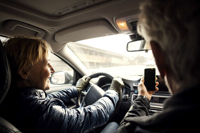 Smiling senior woman driving car while sitting by man taking selfie through smart phone