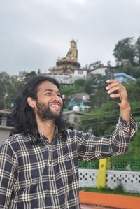 A charming long haired and bearded young guy smiling with taking selfie at outside with mobile phone