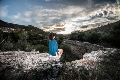 Woman sitting on rock against sky during sunset