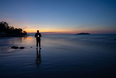 Silhouette man holding camera while standing at beach against sky during sunset
