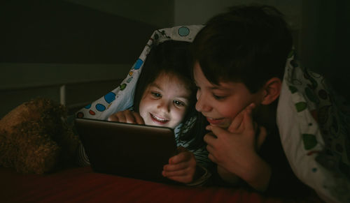 Siblings using digital tablet on bed in darkroom