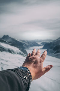 Cropped hand of man gesturing on snowcapped mountain against cloudy sky