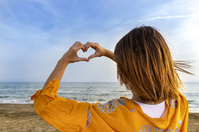 Young woman on the beach making a heart shape with her hands as to declare her love to the sea.