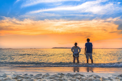 Rear view of friends standing on beach against sky during sunset