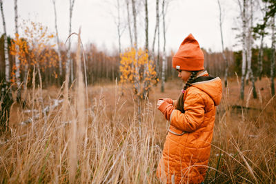 Pensive girl in orange coat stands in forest among dry grass and golden trees, outdoor, fall vibes