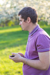 Pleasant cheerful handsome young man holding phone. profile view. nature green garden background.