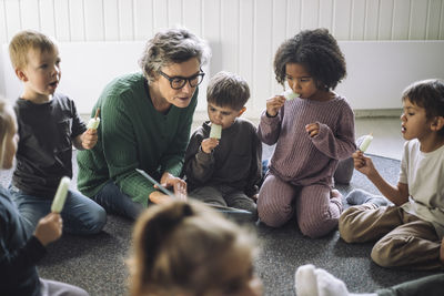 Group of children eating popsicle with senior female teacher while sitting in classroom at kindergarten