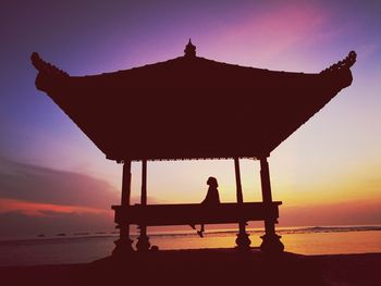 Silhouette woman sitting on bench at beach against sky during sunset