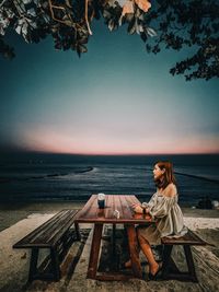 Woman sitting on bench by sea against sky