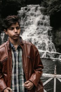 Young man looking away while standing against waterfall