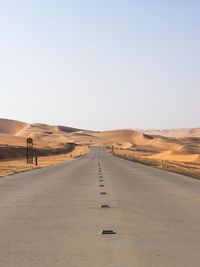 Empty road leading towards sand dunes at desert against clear sky