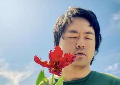 Close-up of young asian man holding red tulip flowering plant against cloudy blue sky.
