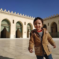 Portrait of smiling cute boy standing on footpath against mosque