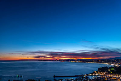 Illuminated city by sea against sky at sunset