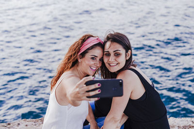 Lesbian couple taking selfie while sitting by sea