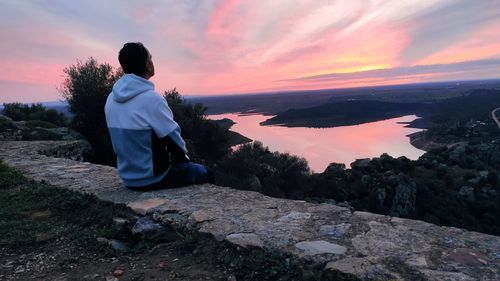 Rear view of man sitting on rock against sky during sunset