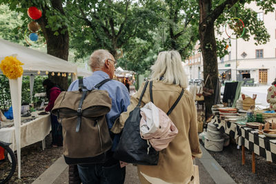 Rear view of senior tourist couple with backpack and bag walking at market in city