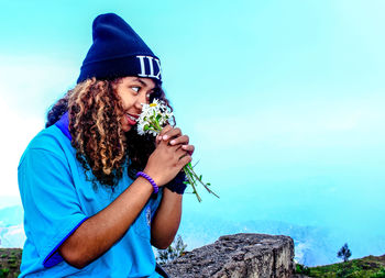 Young woman smelling flowers while standing against sky