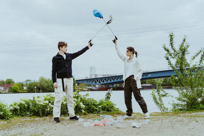 Young friends collecting plastic garbage near river against sky