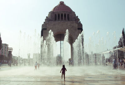 Rear view of girl jumping in front of fountain and historic gate in city
