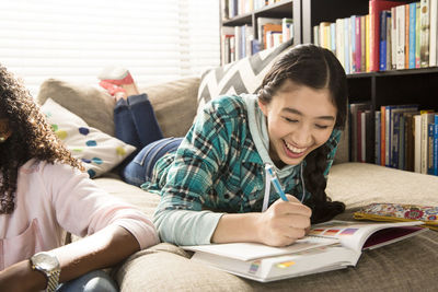 Cheerful teenage girl doing homework while lying on couch by friend at home