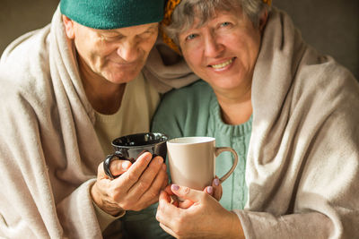 Smiling grandparents in caps wrapped in warm blanket hold cups of hot coffee, feeling cold at home