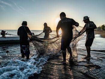People fishing on beach against sky during sunset