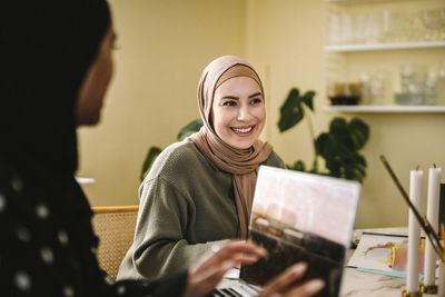 Smiling young woman in hijab sitting by freelancer with laptop at home