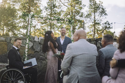 Happy newlywed couple holding bouquet by guests and minister with disability at wedding ceremony