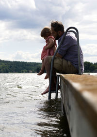 Side view of father carrying daughter while sitting on pier over lake against cloudy sky
