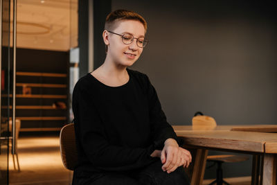 Young woman with short hair in eyeglasses and black sweater sitting at the table in office