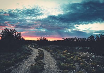 Narrow dirt road on field against cloudy sky during sunset