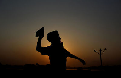 Silhouette man flying paper air plane against sky at sunset