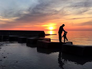 Silhouette father and child walking at beach against sky during sunsets