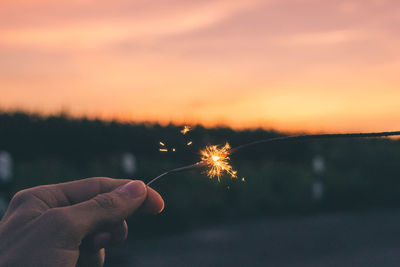 Close-up of hand holding illuminated sparkler against sky during sunset
