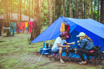 People eating food while sitting by tent in forest