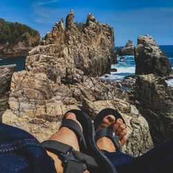 Low section of person wearing sandal sitting on rock by sea