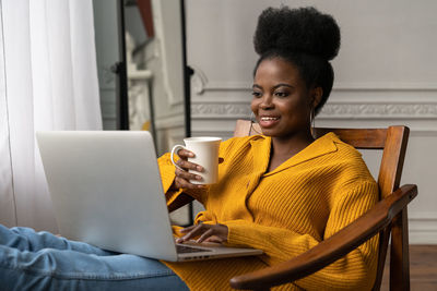 Smiling woman working on laptop while holding coffee cup at home