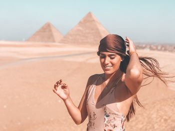 Portrait of smiling woman with her hair on wind  standing on sand in front of pyramids 