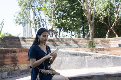 Young woman looking at camera while sitting on retaining wall against trees